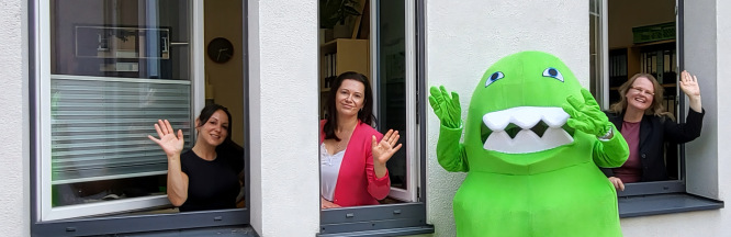 Frau Fenske, Frau Scheller, das Netzwerk-Maskottchen und Frau Dietsch (von links nach rechts) winken am Fenster.